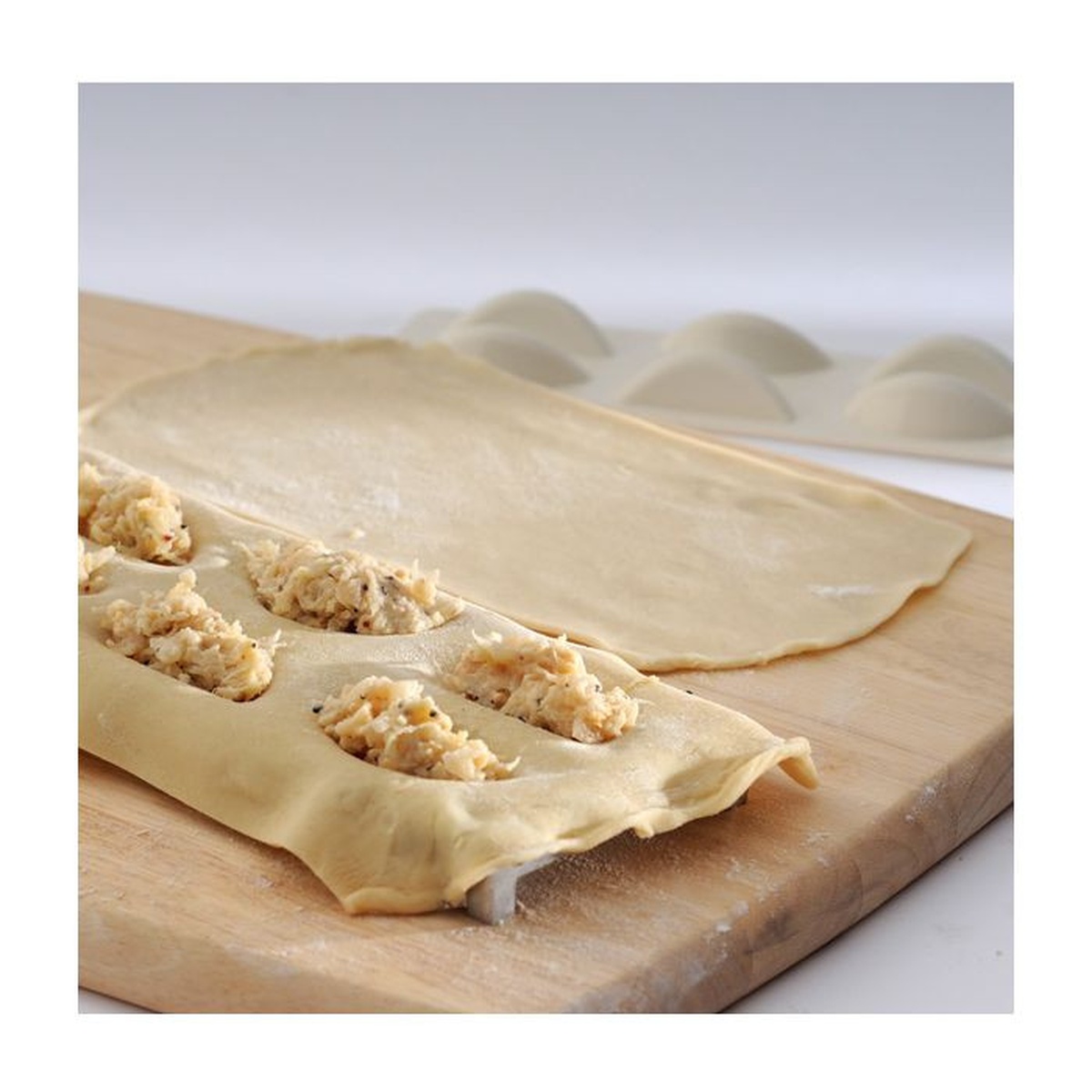 Sheet of pasta dough with savory filling portioned into the pierogi maker cavities on a wooden cutting board, with finished pierogies in the background