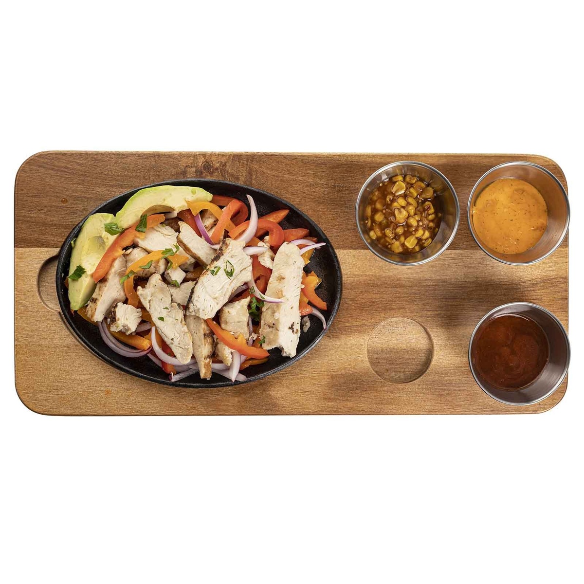 Top-down view of TableCraft 10709 acacia wood fajita board loaded with oval cast iron skillet and three ramekins filled with sauces