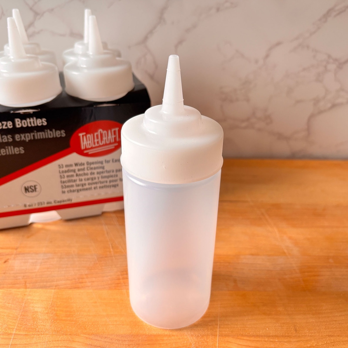 TableCraft 10853C clear widemouth squeeze bottle standing on a wooden cutting board beside TableCraft branded squeeze bottle packaging