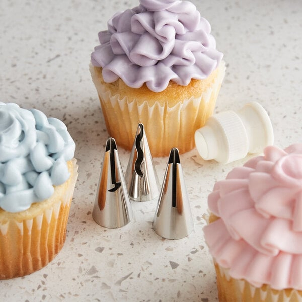 Three Ateco ruffle piping tips and a plastic coupler displayed alongside decorated cupcakes showing purple, blue, and pink ruffle frosting designs