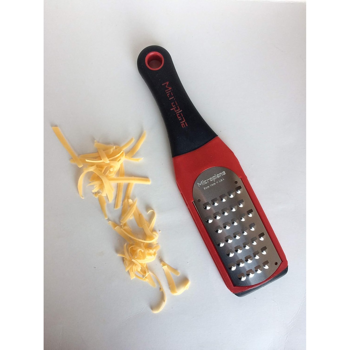 Microplane Artisan Extra Coarse Grater in red next to a pile of freshly shredded cheddar cheese on a white surface