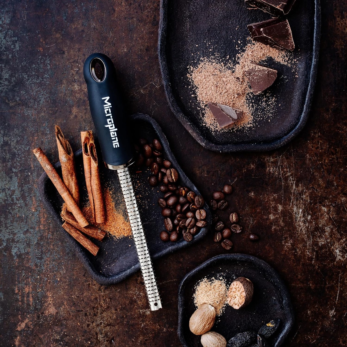 Microplane spice grater on a dark surface with cinnamon sticks, coffee beans, and chocolate