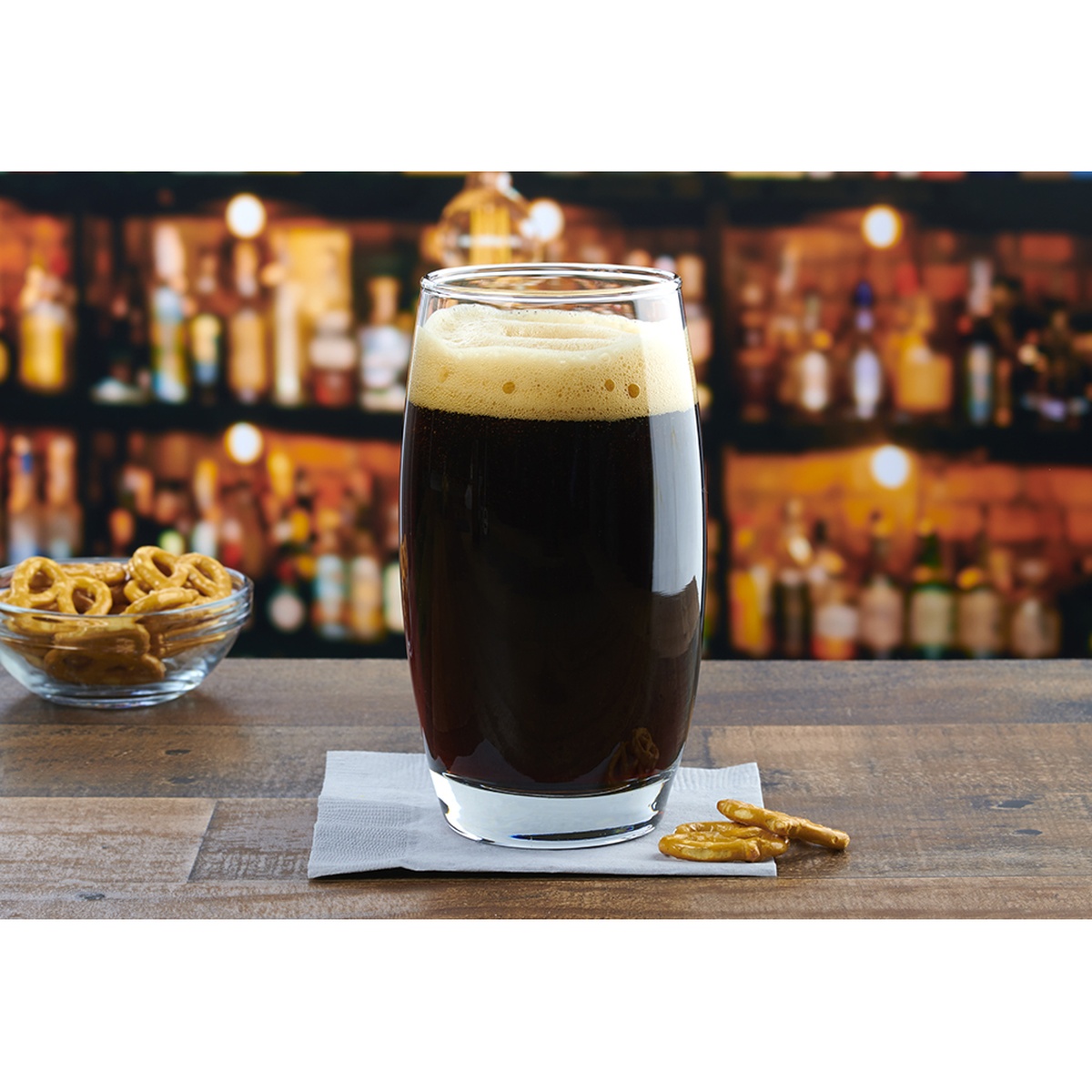 Anchor Hocking Reality 20 oz. cooler glass filled with dark stout beer, displayed on a wooden bar surface with a bowl of pretzels in the background