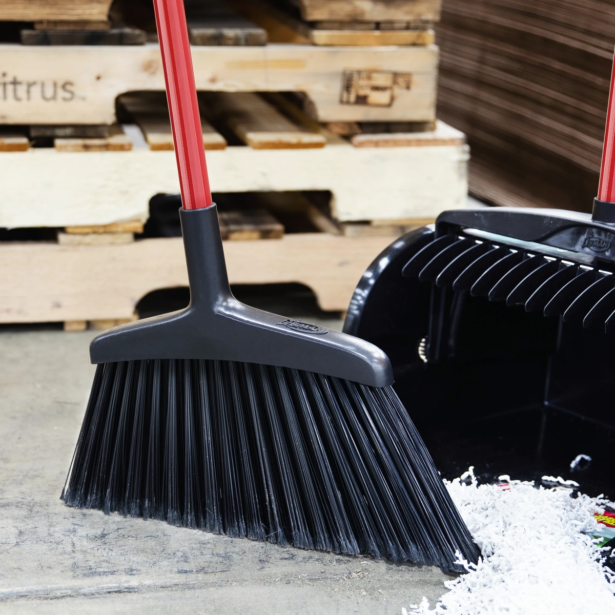 Libman 997 wide commercial angle broom with red handle sweeping shredded paper debris into a dustpan on a concrete floor