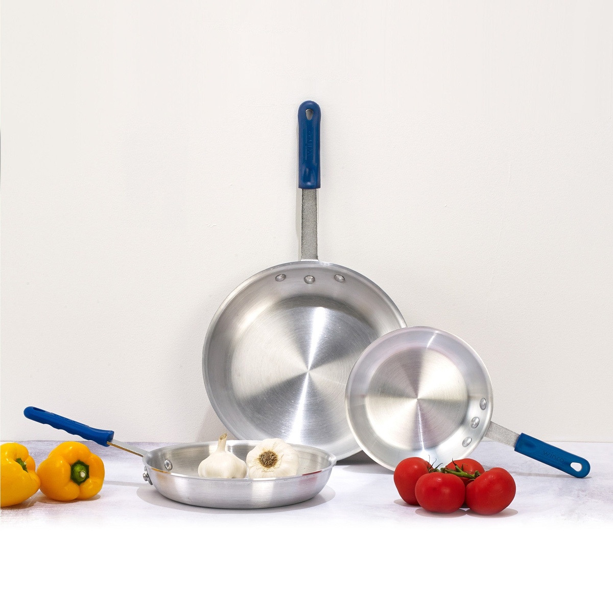 Winco blue silicone sleeves shown on various sized aluminum fry pans with fresh vegetables