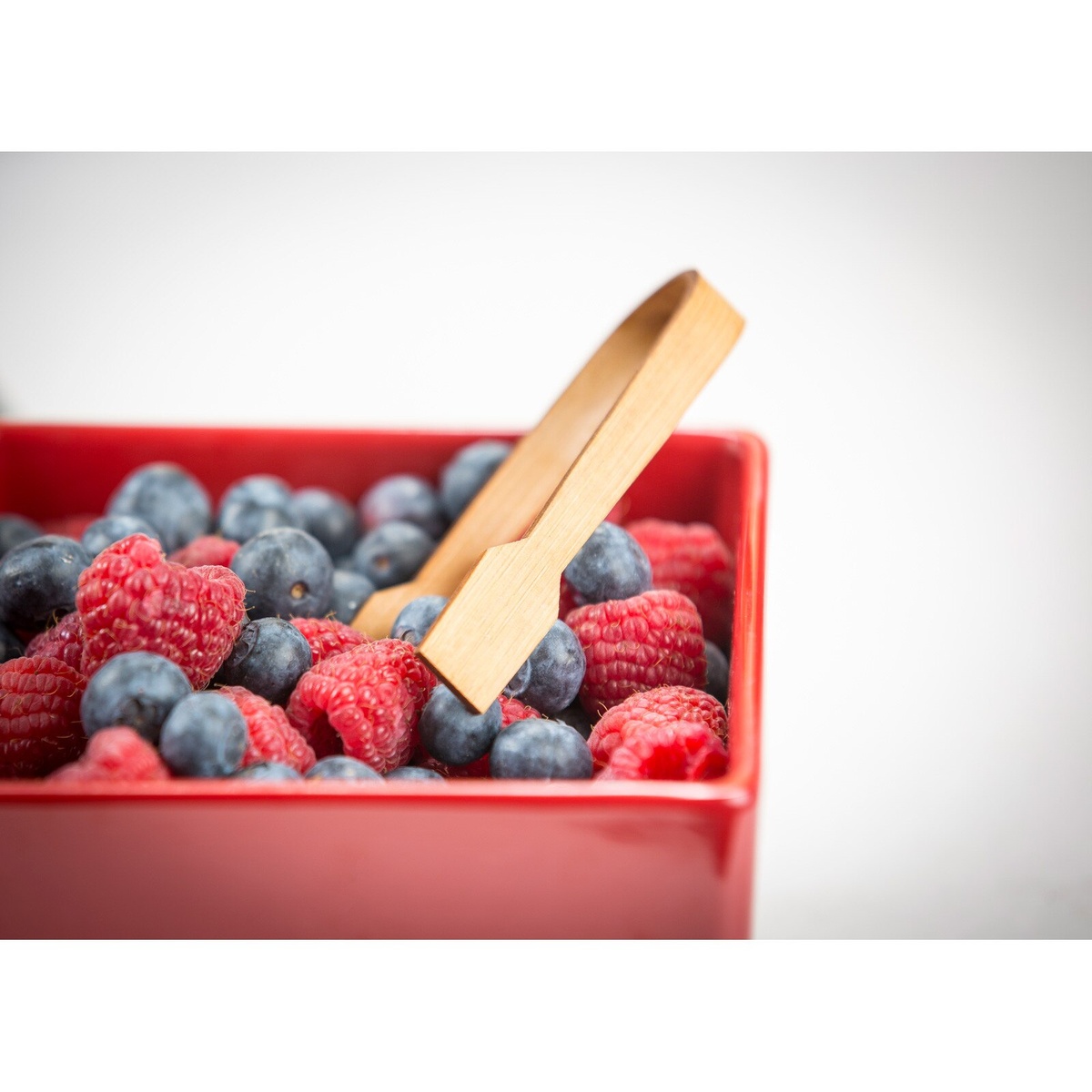 Bamboo paddle tongs being used to pick up fresh berries in a red bowl