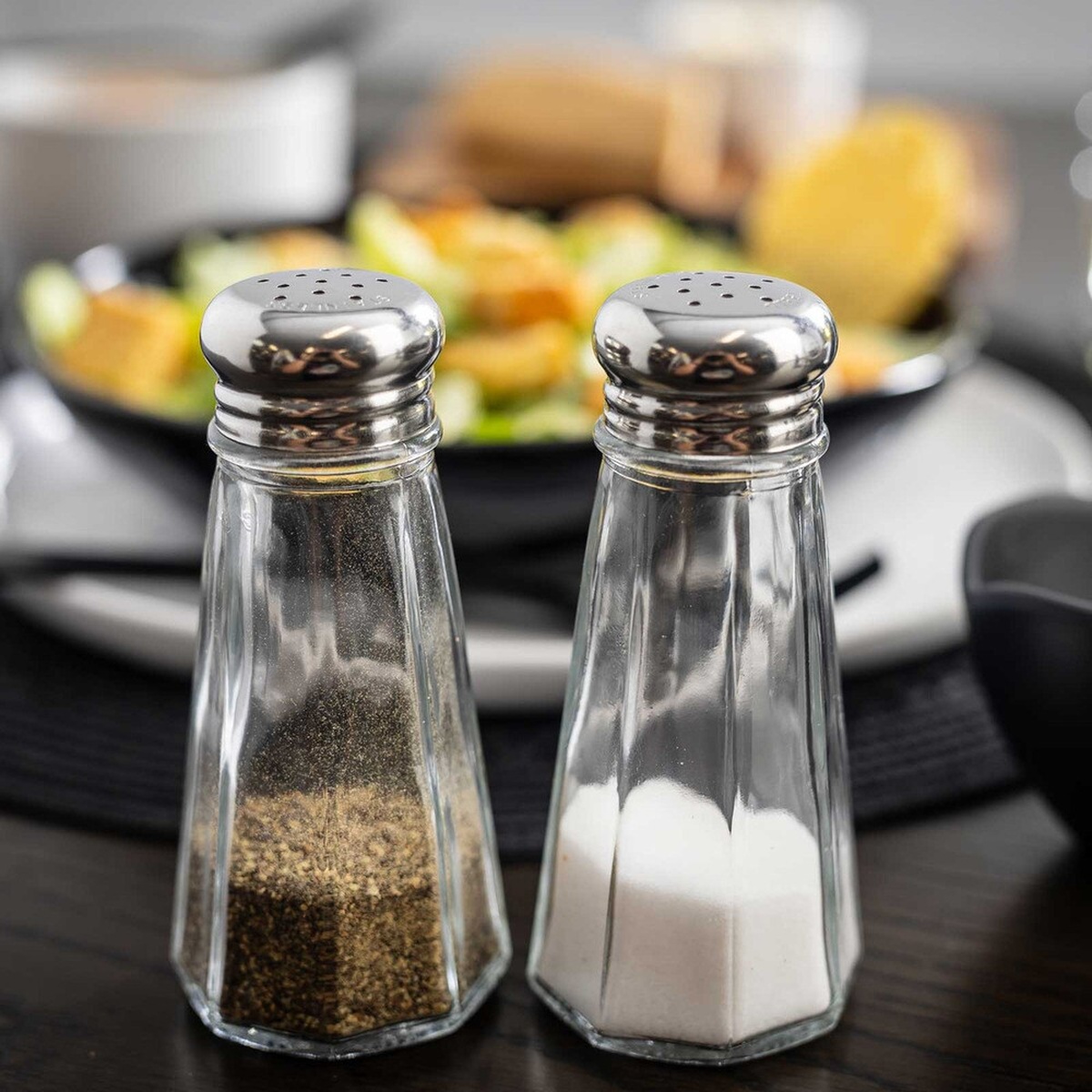 Two TableCraft C156-12 paneled glass salt and pepper shakers with stainless steel tops on a dining table
