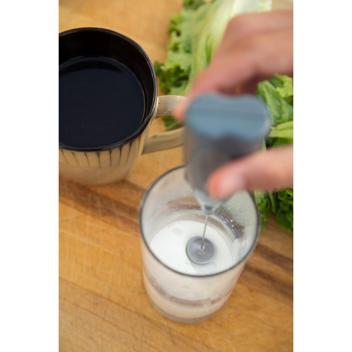 Milk frother frothing milk in a glass next to a coffee mug on a wooden surface