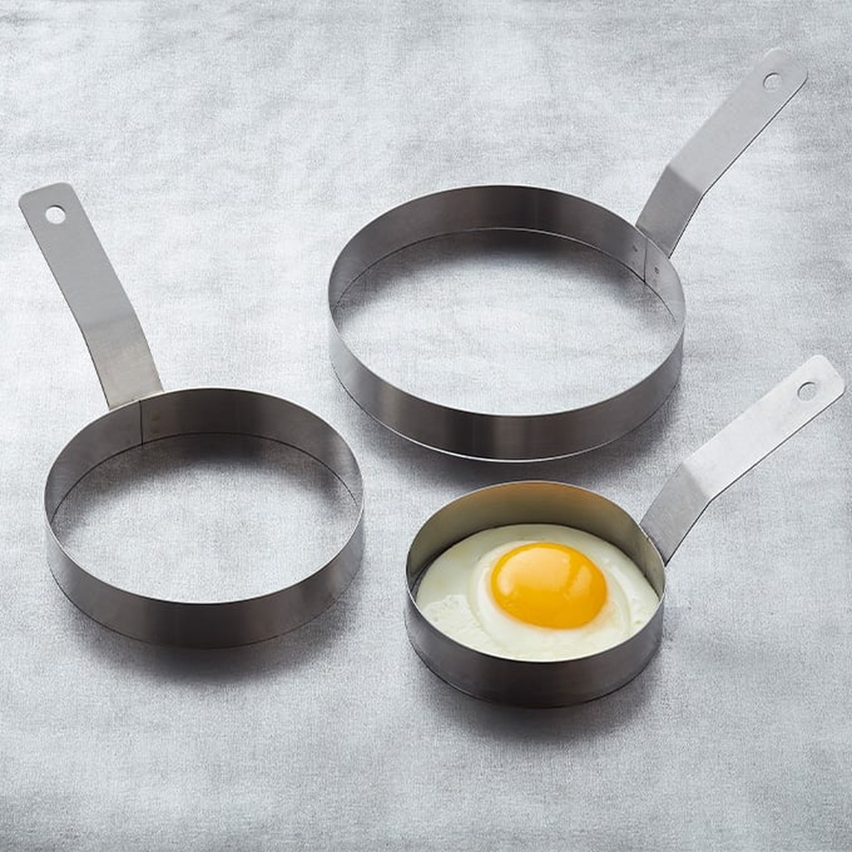 Three American Metalcraft stainless steel egg rings of different sizes displayed on a gray surface, one containing a fried egg