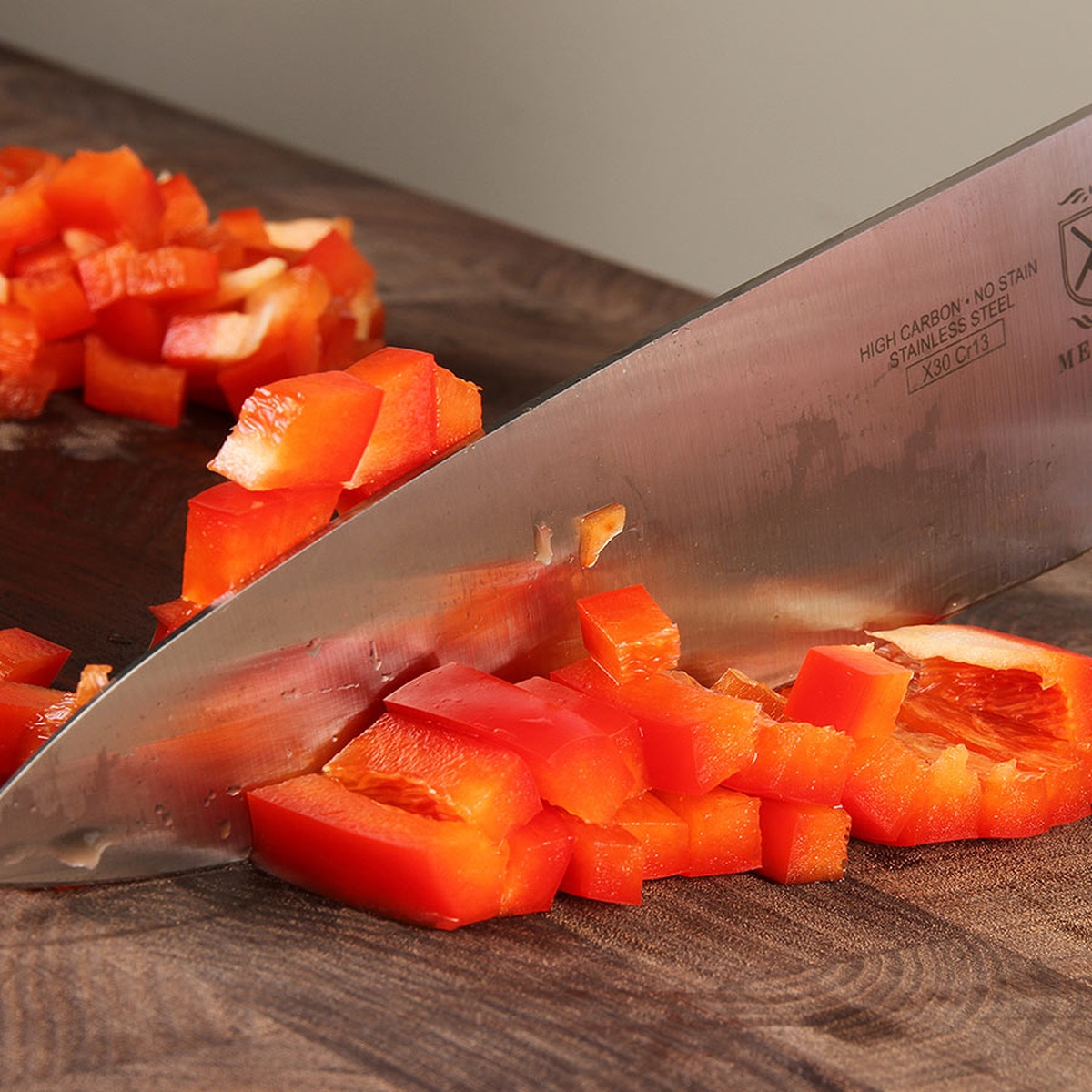 Close-up of Mercer Culinary chef's knife blade slicing diced red bell peppers on a wooden cutting board