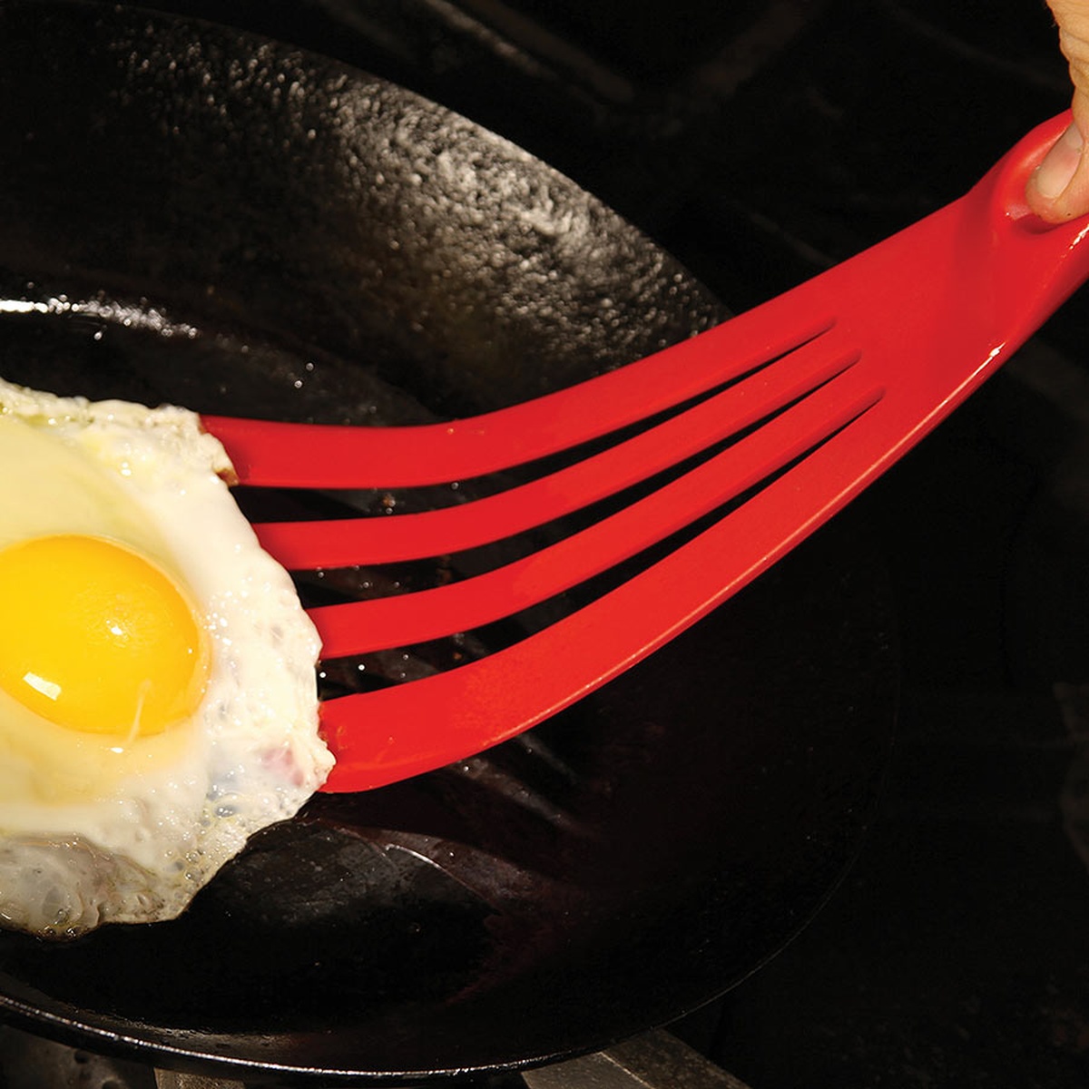 Red slotted spatula flipping a fried egg in a cast iron skillet