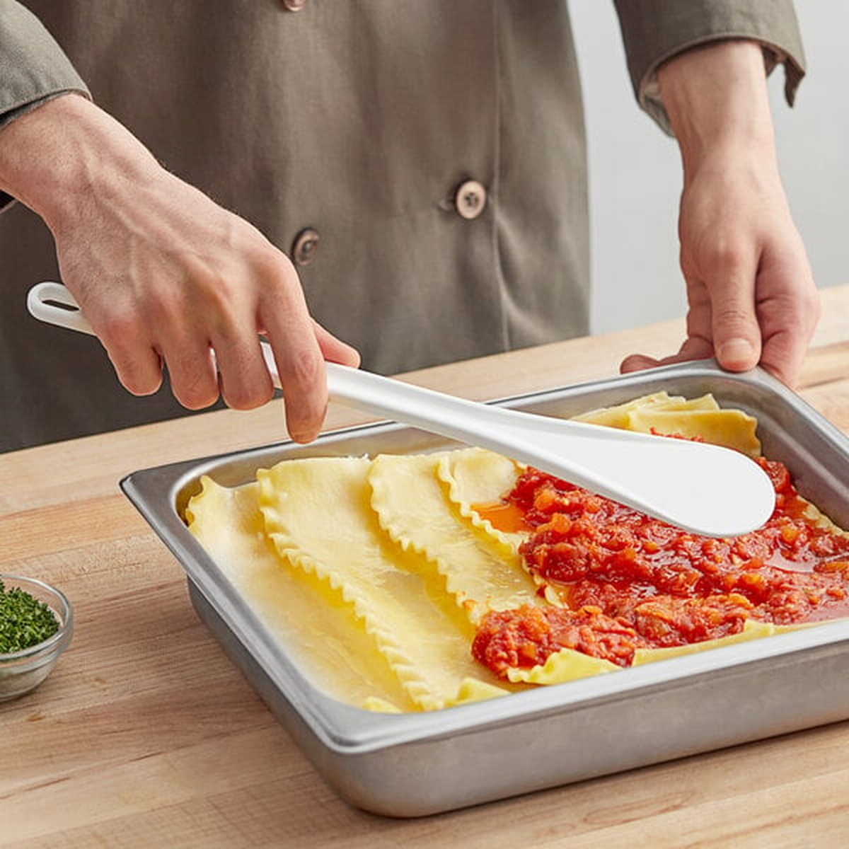 Chef using a white spootensil to spread tomato sauce over lasagna noodles in a metal pan