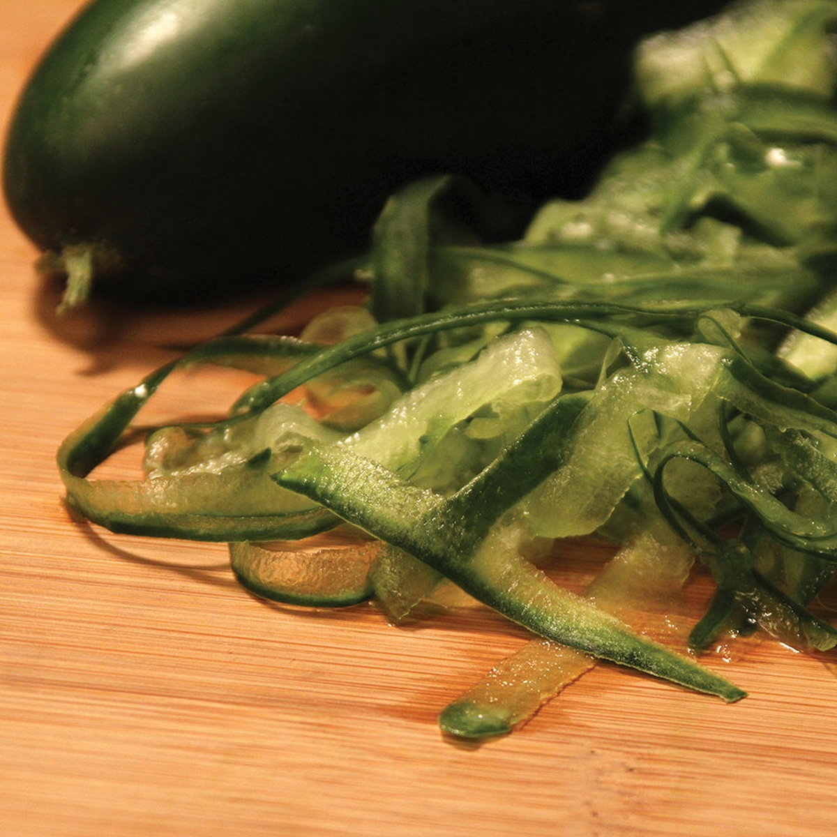 Fresh zucchini ribbons on a wooden cutting board next to a whole zucchini, demonstrating the output of the MercerGrates ribbon grater