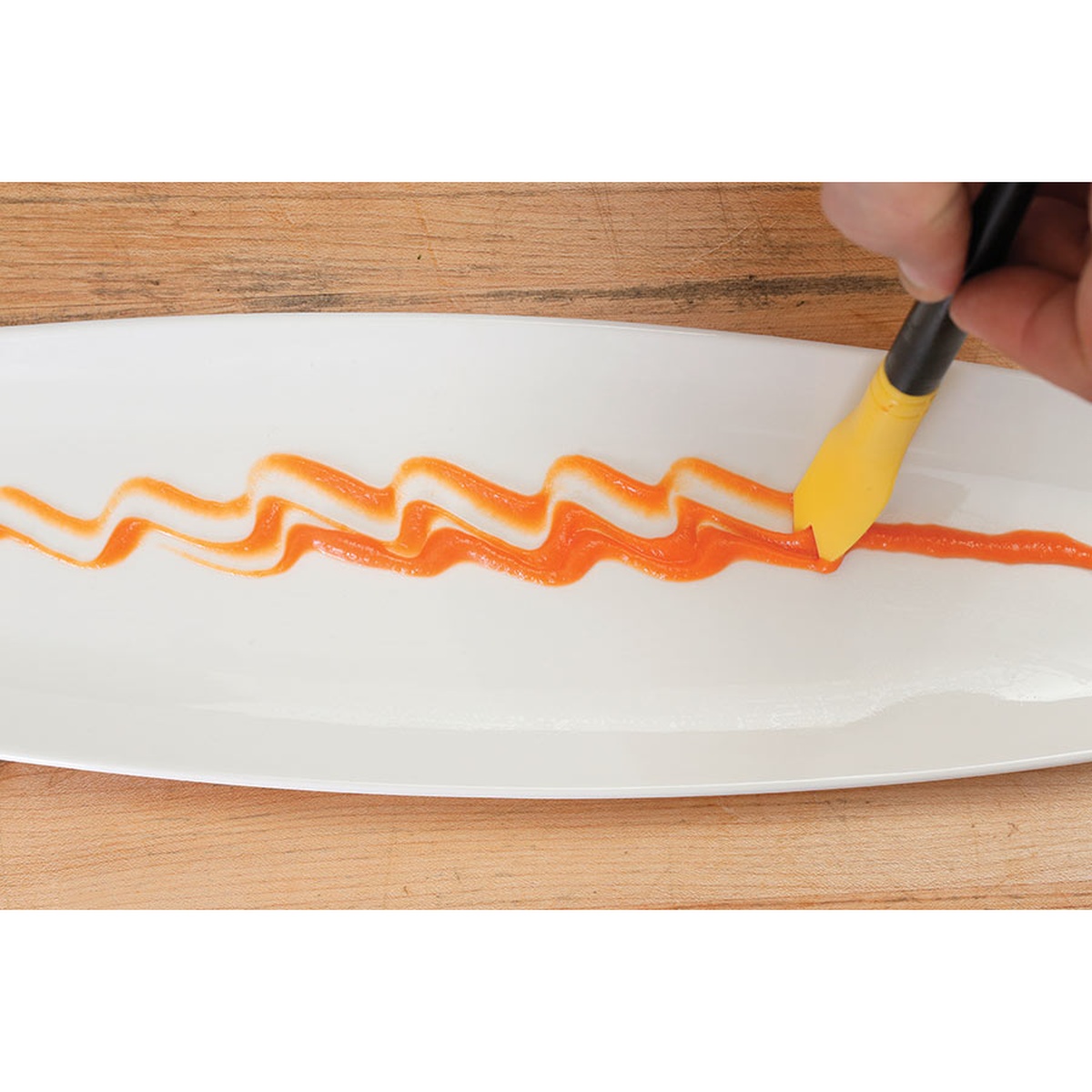 Close up of the silicone plating tool creating a wavy orange sauce pattern on a long white dish