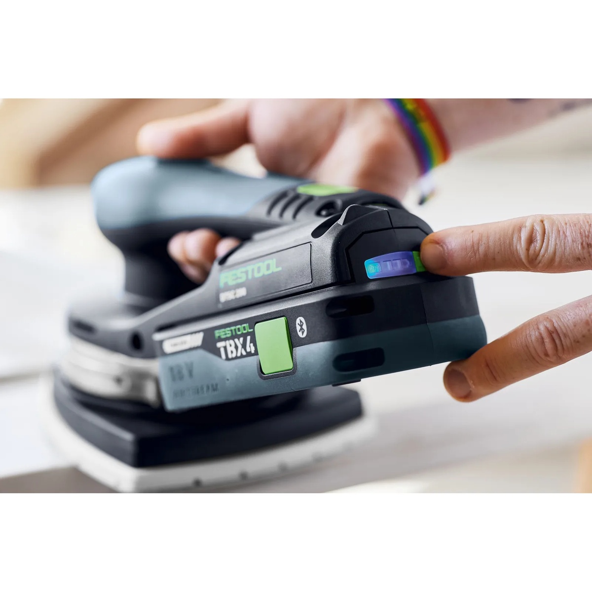 Festool BP 18 TBX 4 C-ASI battery pack installed on a cordless sander while a user checks the battery status indicator during finish sanding on a work surface
