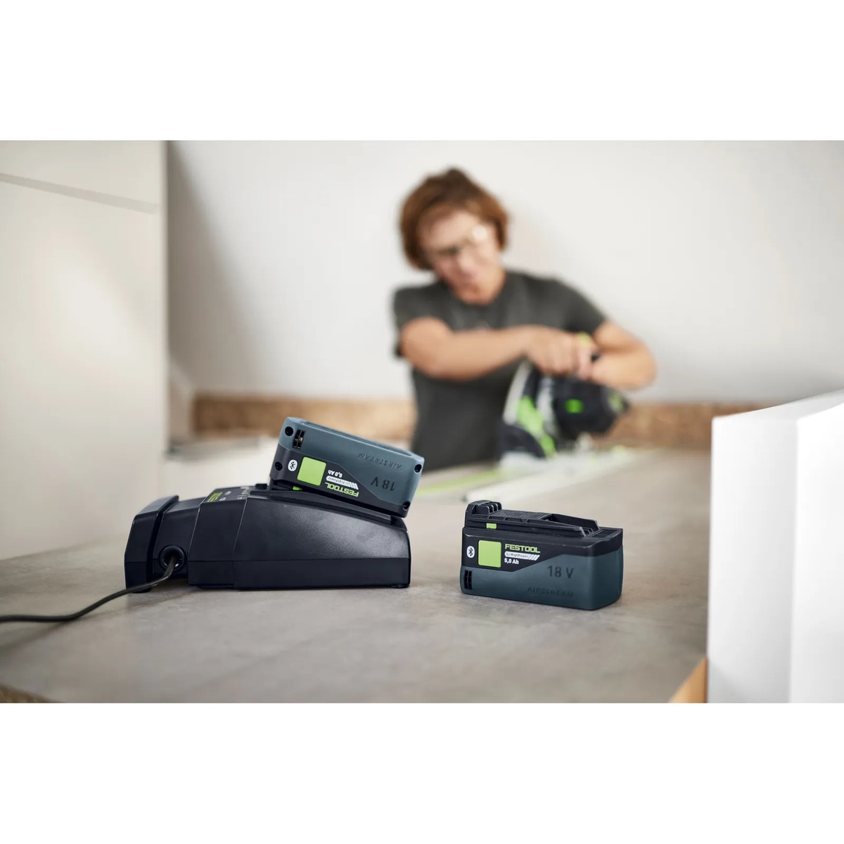 Festool BP 18 Li 5,0 HP-ASI HighPower battery pack charging on a Festool charger on a work surface while a user works with a Festool cordless tool in the background