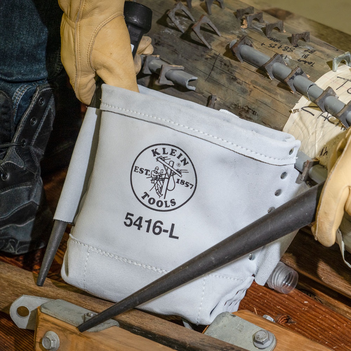 Klein Tools Tool Bag, Bull-Pin and Bolt Bag, Tunnel Loop, Leather, 5 x 10 x 9-Inch holding steelworking hardware on a work surface with gloves and bull pins during structural fastening work