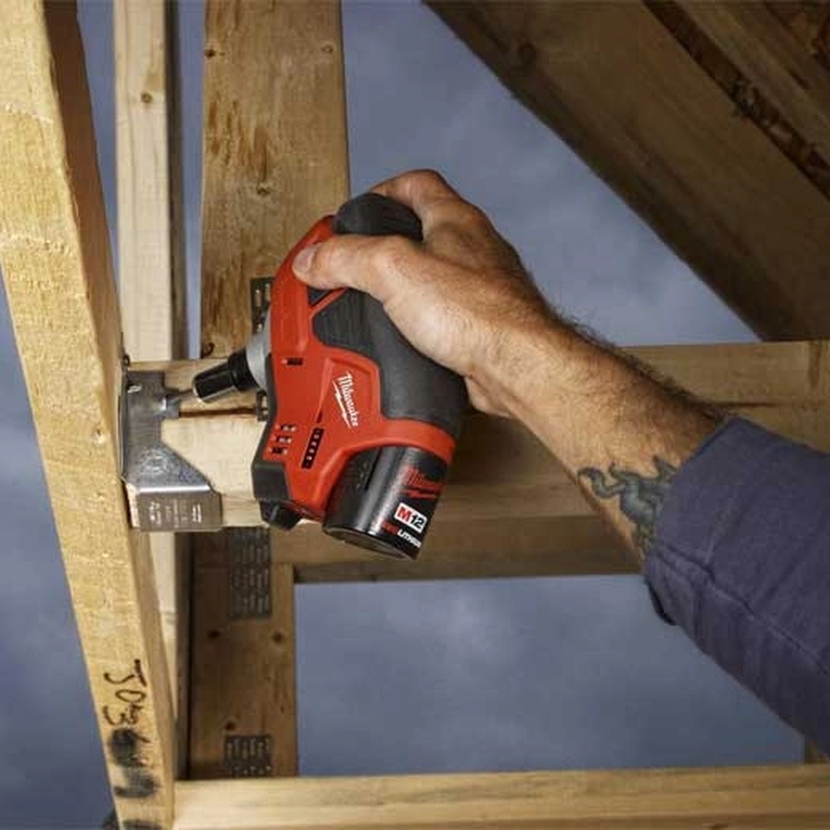 Milwaukee 2458-20 M12 Cordless Palm Nailer driving a nail into framing lumber beside a metal connector in a tight overhead framing bay