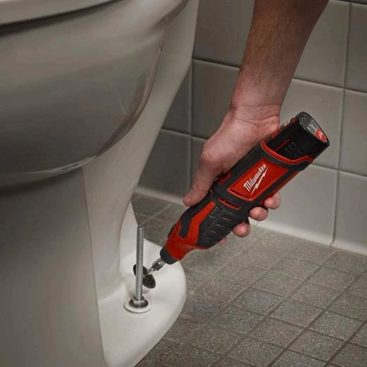 Milwaukee M12 Cordless Lithium-Ion Rotary Tool grinding fastener hardware near a toilet base during plumbing and repair work