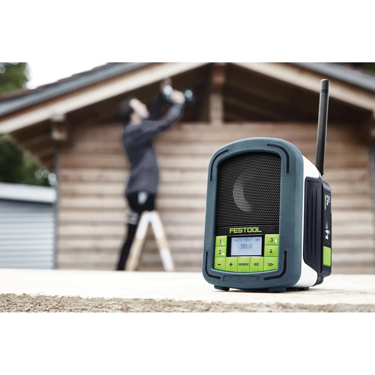 Festool 200184 BR10 SysRock Jobsite Radio set on a work surface outdoors at a jobsite while a worker is on a ladder in front of a wood building