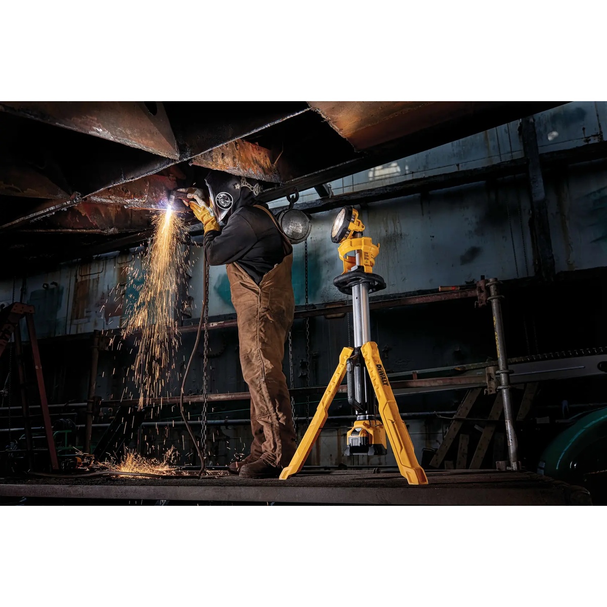 DEWALT DCL079R1 20V MAX LED work light and tripod kit illuminating a dark industrial work area while a worker performs overhead metal work