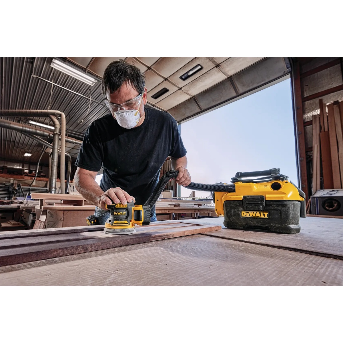 DEWALT DCW210B random orbital sander connected to dust extraction while sanding wood boards on a workbench in a woodworking shop