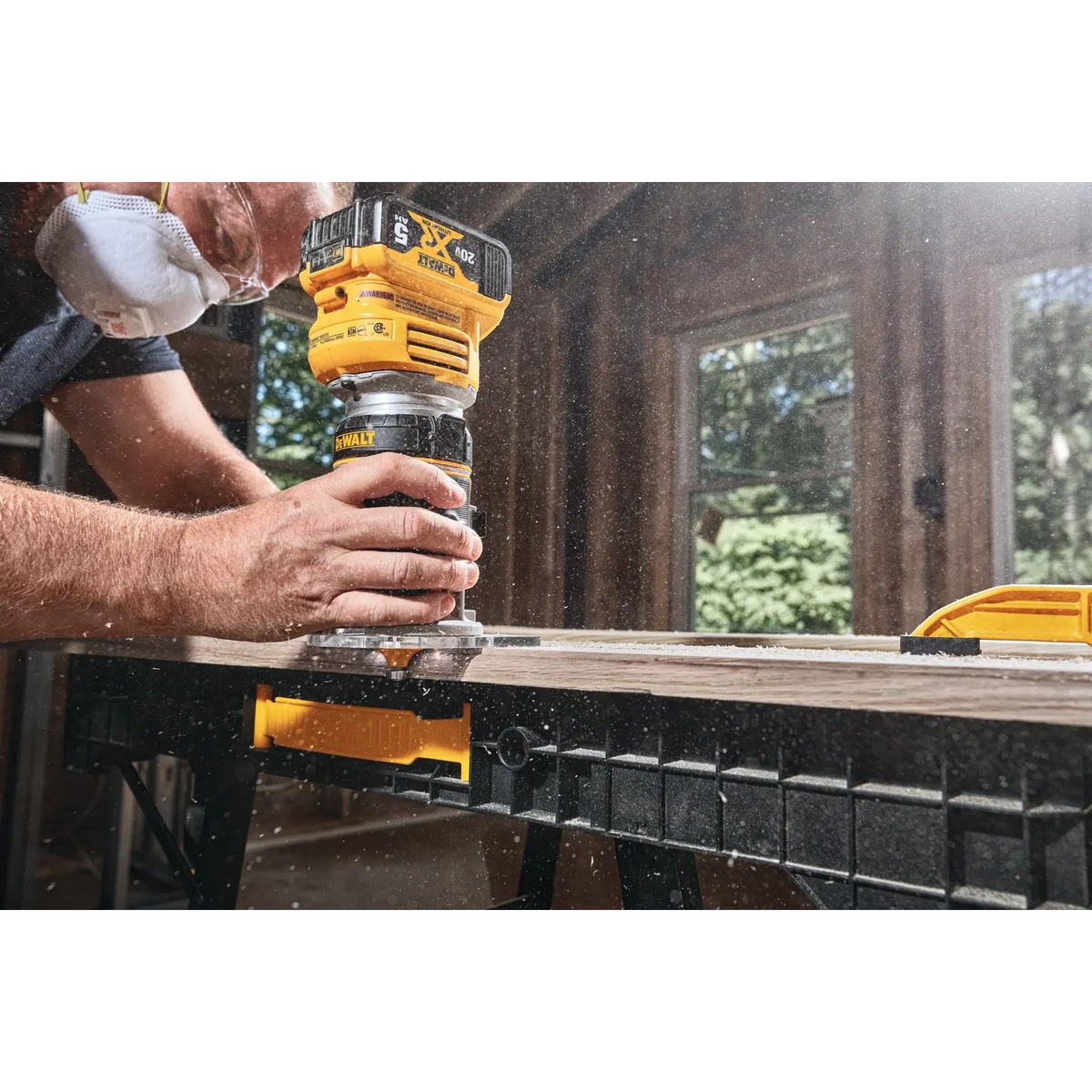 DEWALT DCW600B 20V MAX XR Brushless Cordless Router trimming a wood board on a workbench during indoor woodworking with visible dust in the air