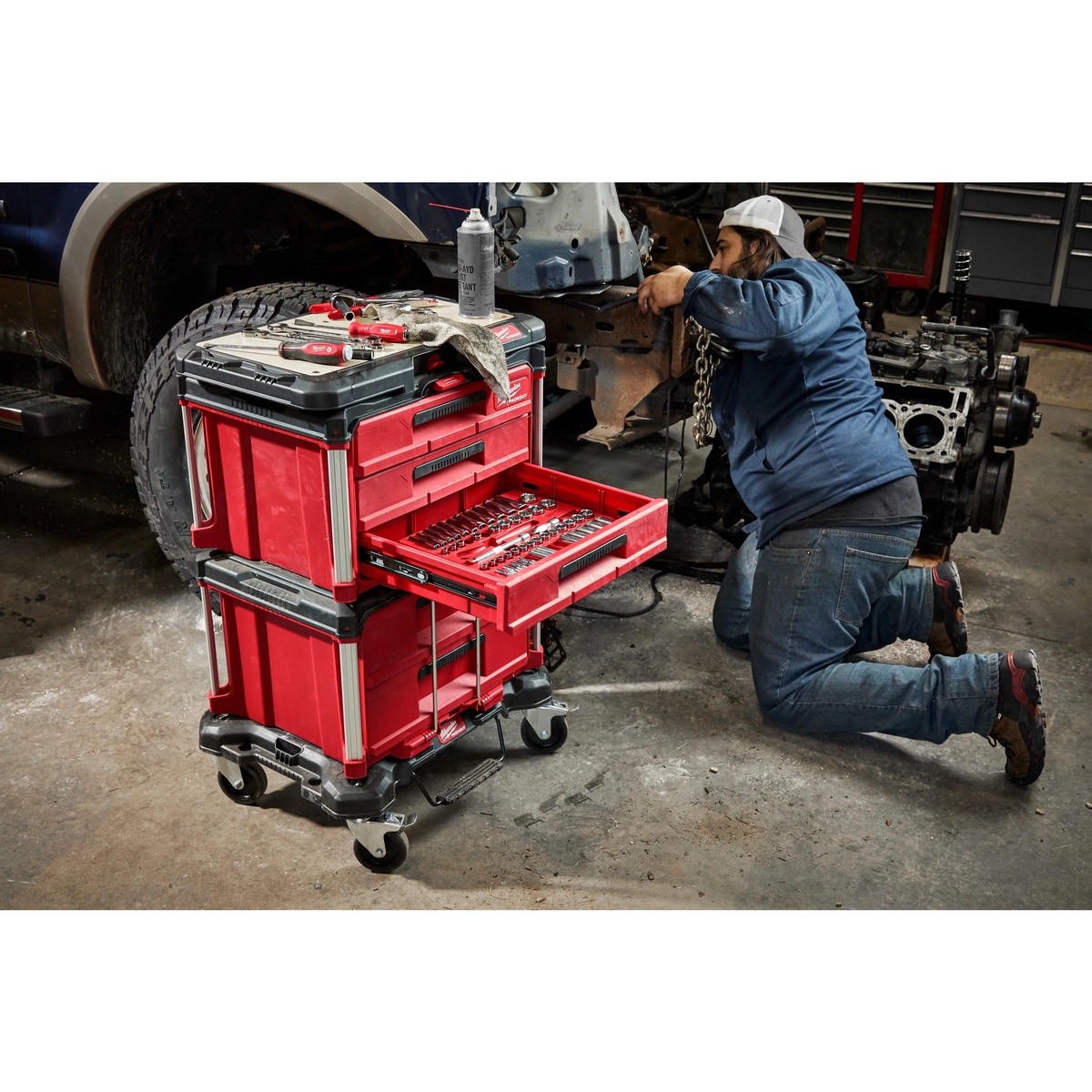 Milwaukee PACKOUT Dolly supporting a stacked PACKOUT drawer and tool box setup in an automotive shop beside a vehicle during repair work