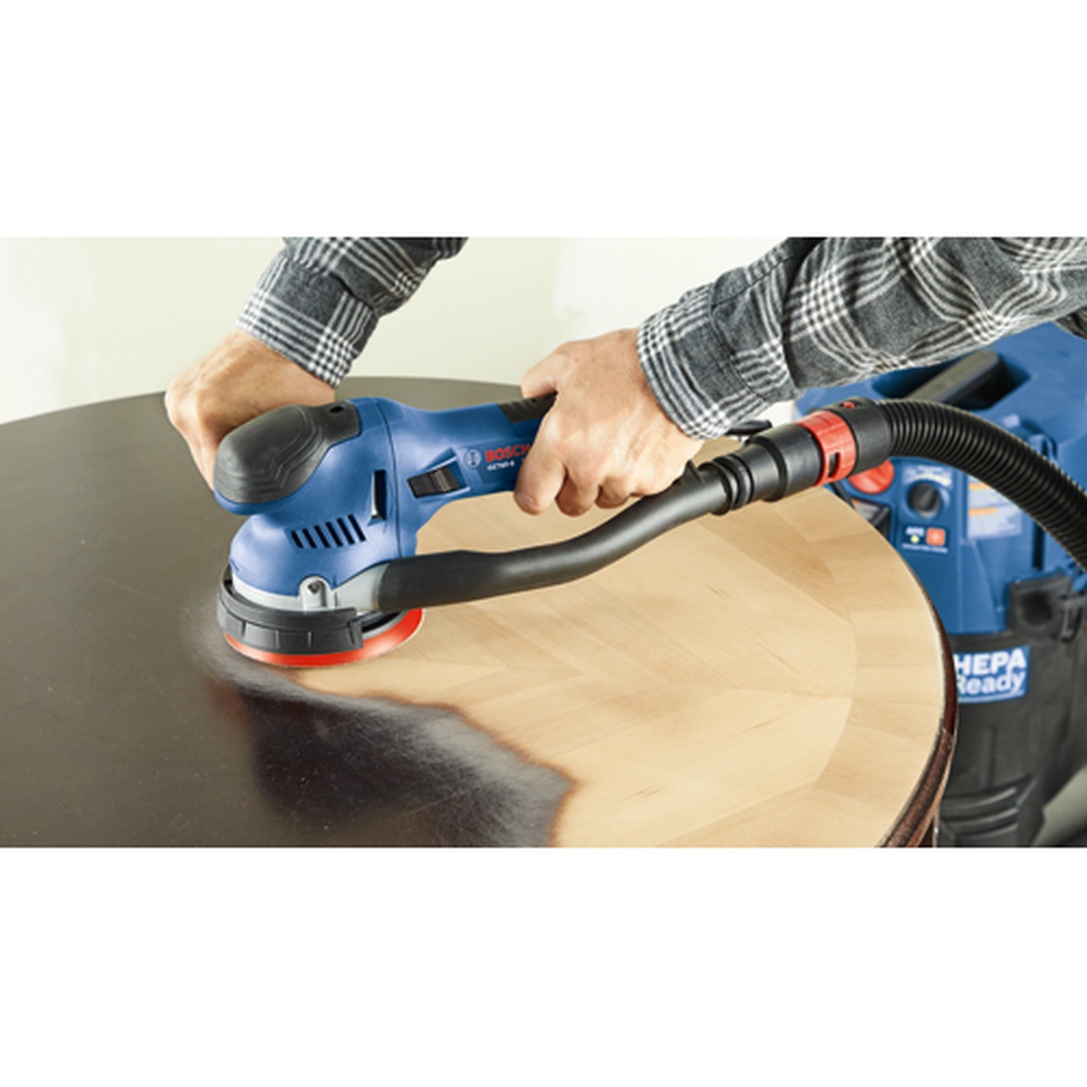 Bosch GET65-5N 5 In. Dual-Mode Random Orbit Sander stripping finish from a round wood tabletop while connected to a vacuum in a workshop setting