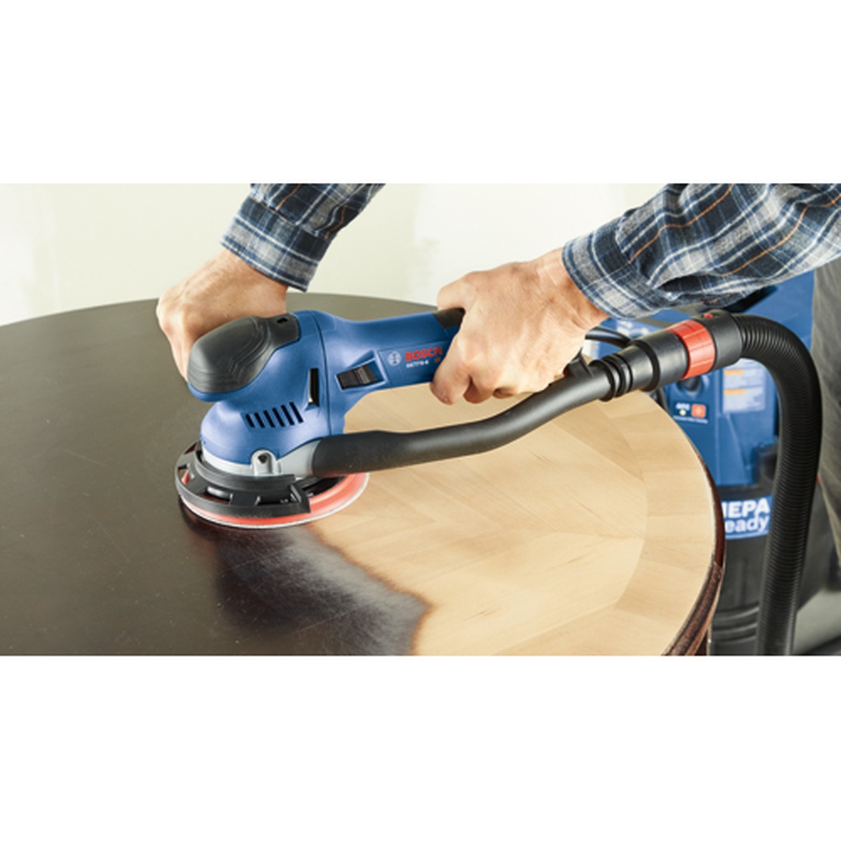 Bosch GET75-6N 6 In. Dual-Mode Random Orbit Sander sanding a round wood tabletop while connected to a dust extractor in a shop setting
