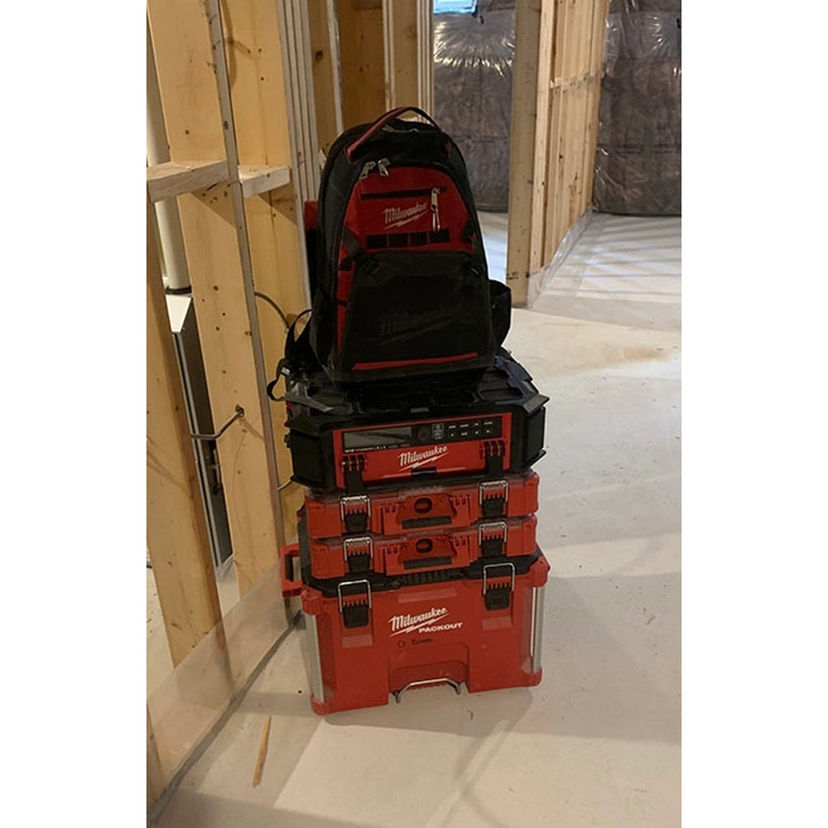 Milwaukee M18 Jobsite Radio + Charger w/ PACKOUT Compatibility stacked in a framed interior jobsite with PACKOUT storage and backpack on top