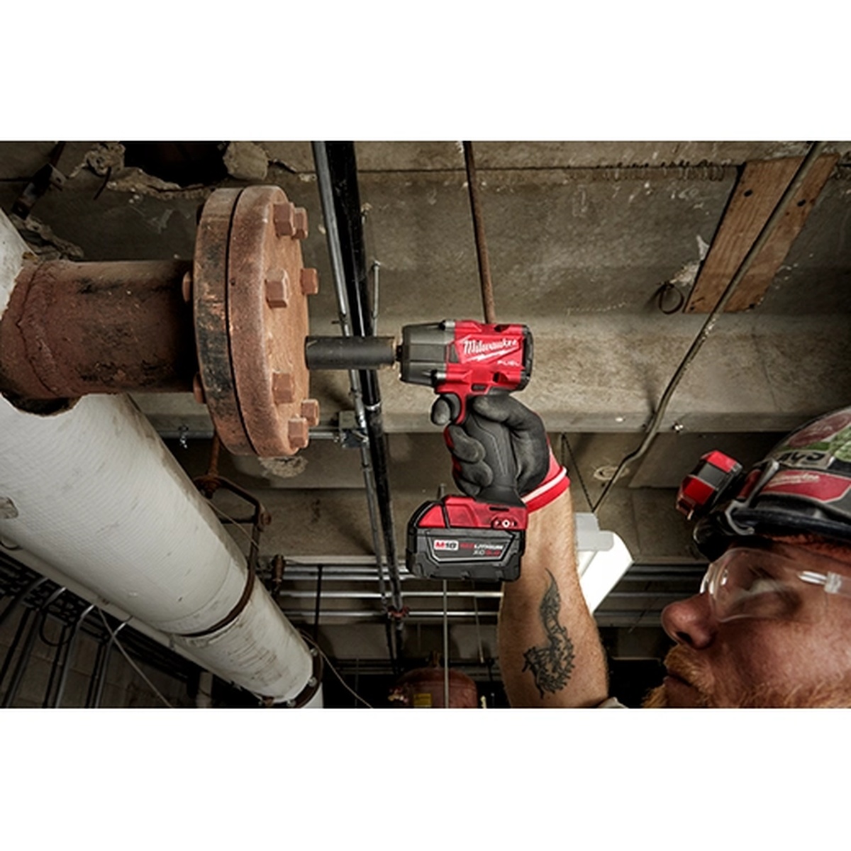 Milwaukee 2962P-20 M18 FUEL 1/2 inch mid-torque impact wrench removing a large fastener overhead on industrial piping in a mechanical room