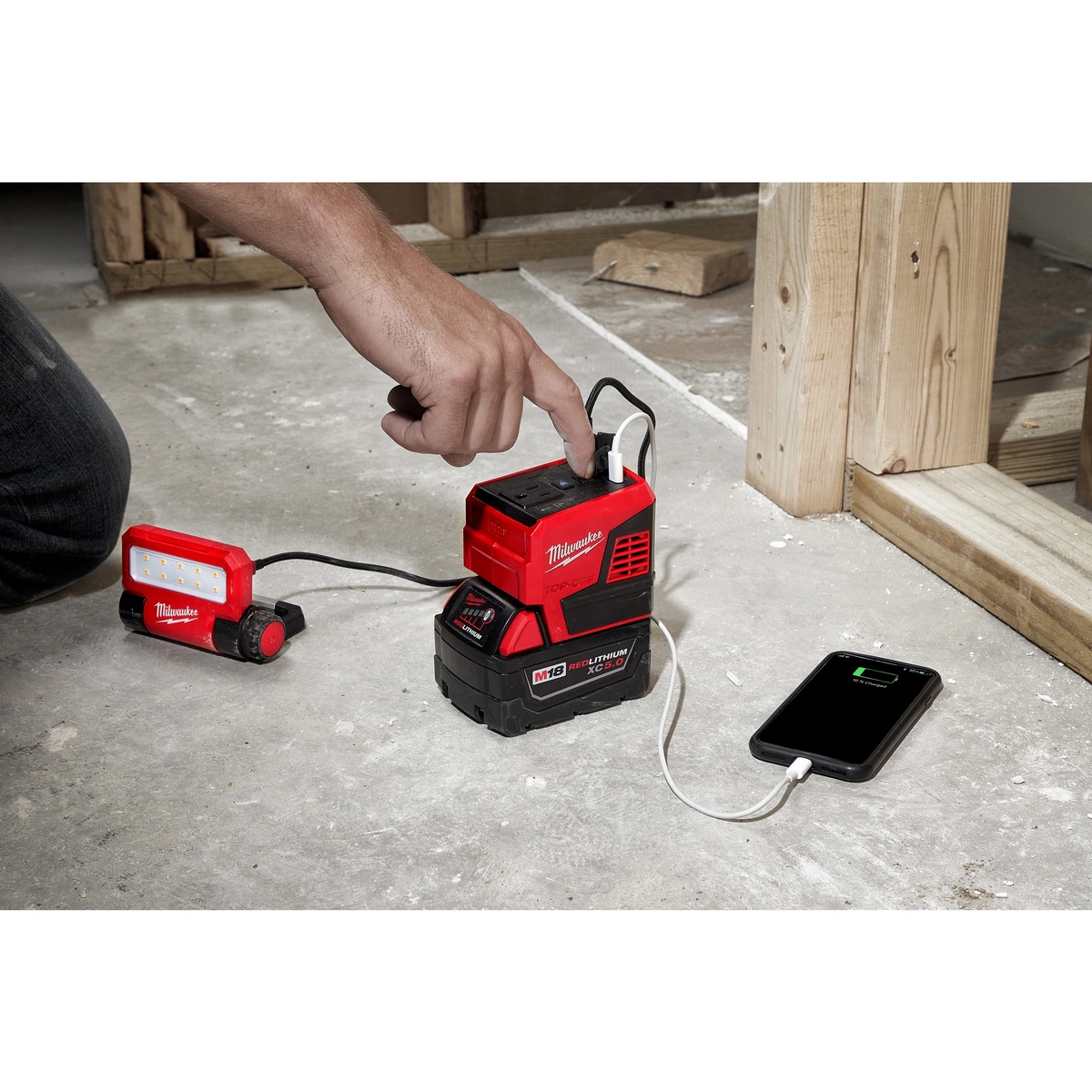 Milwaukee M18 TOP-OFF 175W Power Supply charging a phone and powering a Milwaukee work light on a jobsite floor near framing lumber
