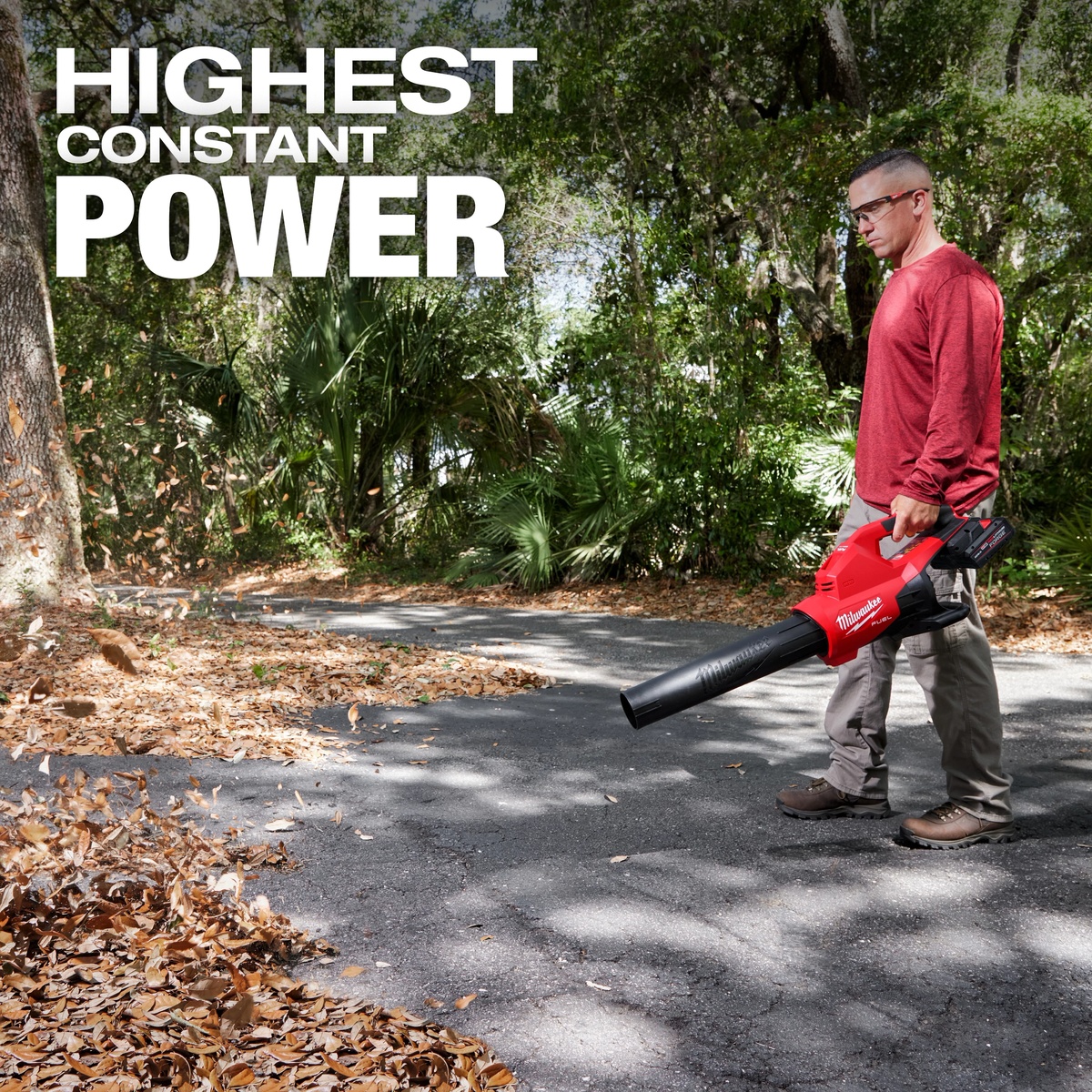 Milwaukee M18 FUEL Dual Battery Blower clearing dry leaves from pavement in a wooded outdoor property maintenance setting