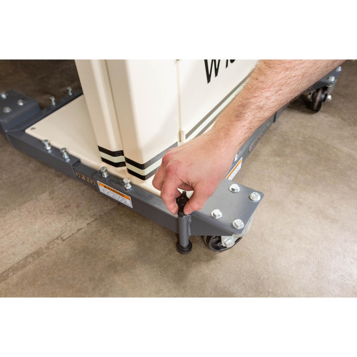 Shop Fox D2058A Adjustable Mobile Base retractable foot being adjusted by hand beside a mounted machine on a workshop floor