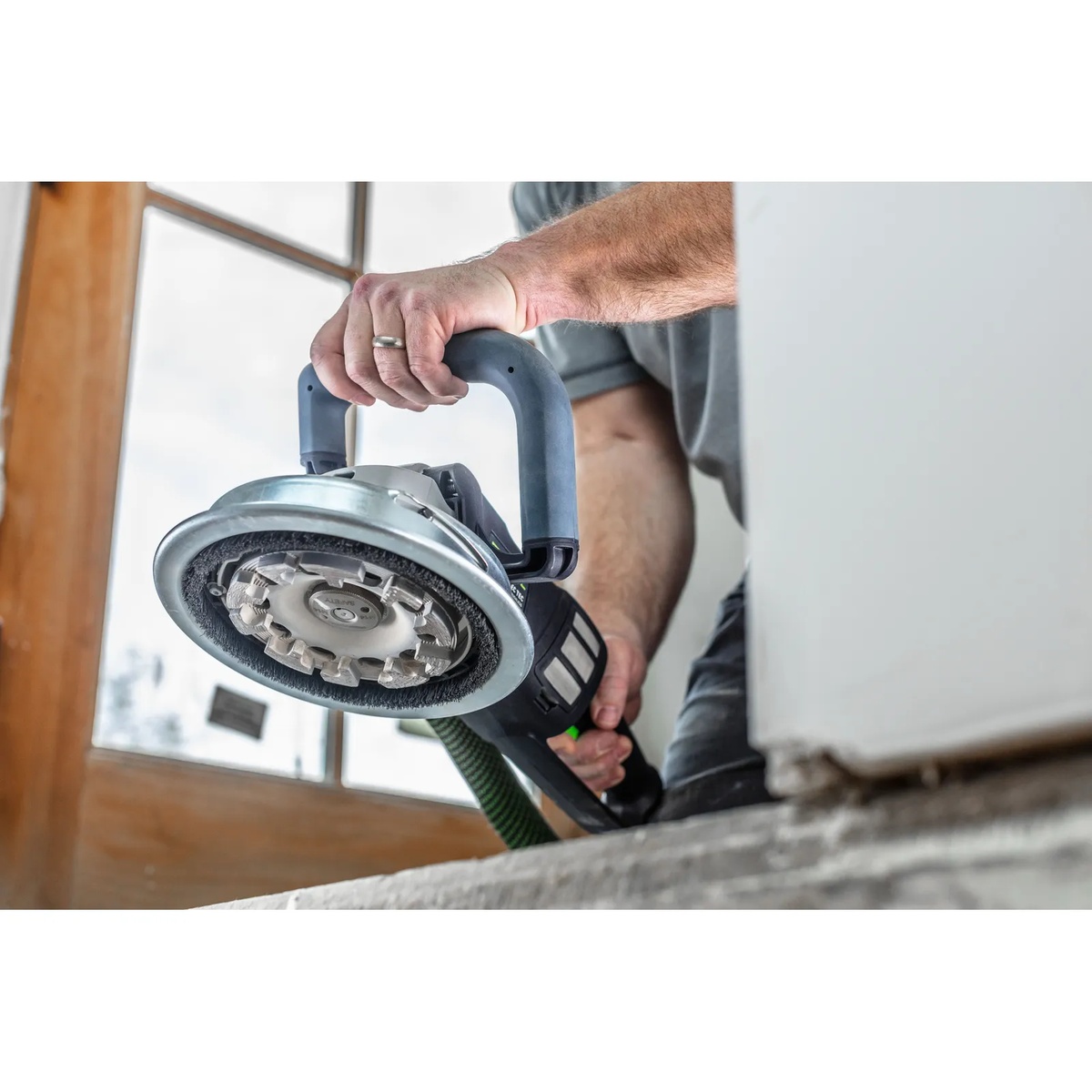 Festool 577048 Corded RENOFIX RG 130 ECI-Plus Surface Restoration Grinder Set showing the grinding head and edge access near a wall during concrete prep