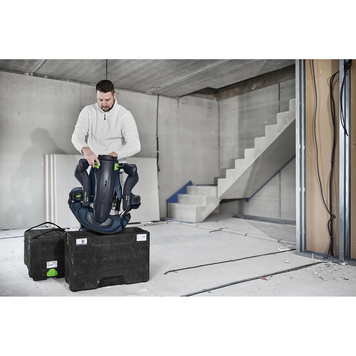 Festool ExoActive exoskeleton being set up on a jobsite floor beside transport cases in a concrete interior during prep for wall and ceiling finishing work