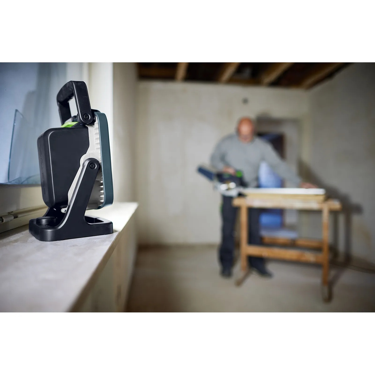 Festool SYSLITE KBS C cordless jobsite light on a windowsill lighting an indoor workspace while a contractor works in the background