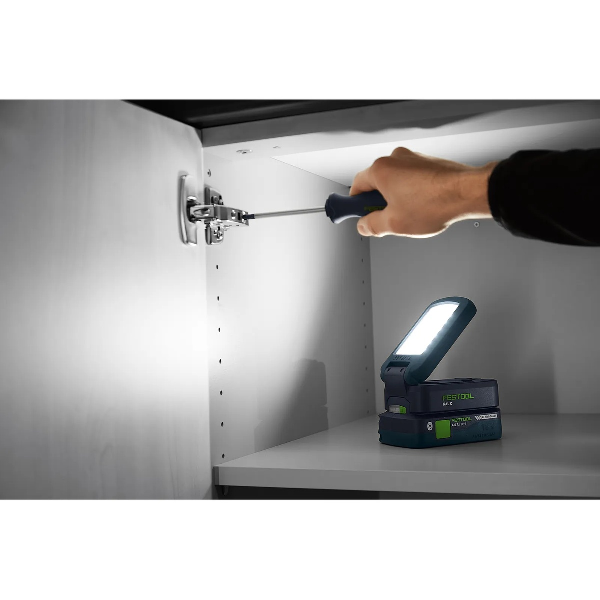 Festool 578128 SYSLITE KAL C Cordless 1200-Lumen Task LED Light illuminating the inside of a cabinet while a user adjusts cabinet hardware in a tight installation space