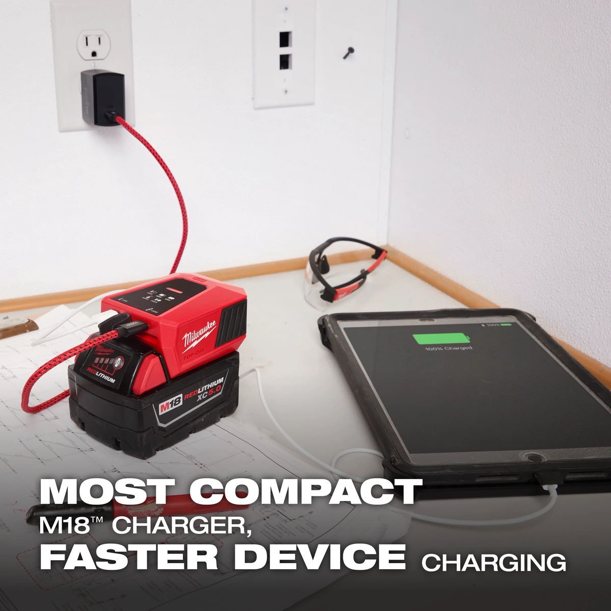 Milwaukee 2847-20 M18 TOP-OFF Charger & Power Supply charging a tablet from an M18 battery on a work surface near plans and safety glasses