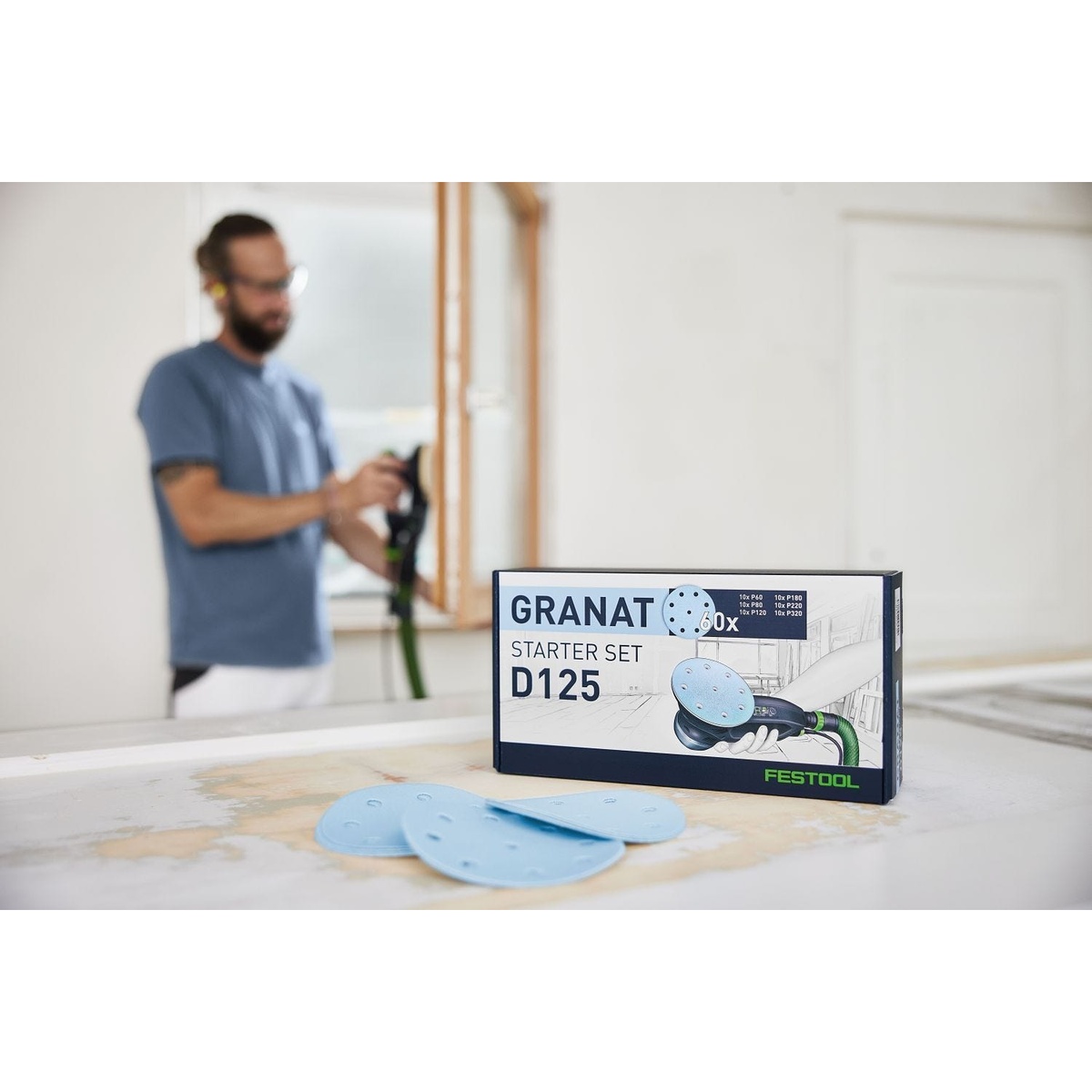 Festool Cordless 18V ETSC 2 125mm (5-in.) Sander Kit w/ 60-Piece GRANAT Abrasive Starter Pack box and sanding discs on a benchtop in a workshop with sanding work in the background