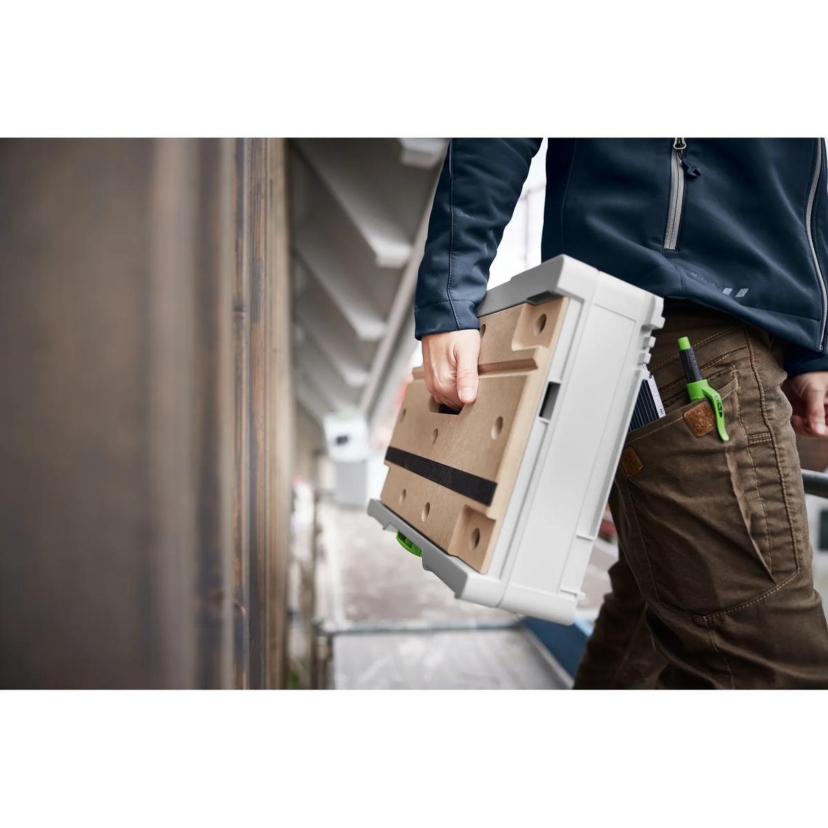 Festool SYS3-MFT M 112 Systainer being carried by hand on a jobsite walkway, showing the portable workbench case ready for trim and fitting work
