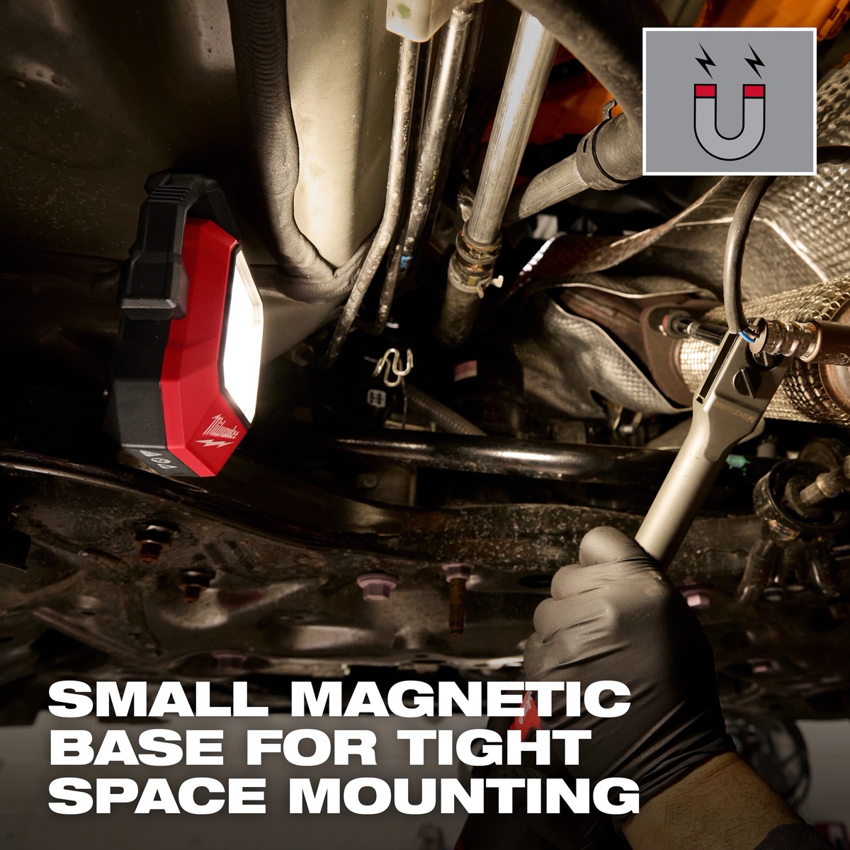 Milwaukee Rechargeable Pivoting Task Light w/ Spot Mode mounted under a vehicle with its magnetic base, lighting an automotive repair area in a tight service bay