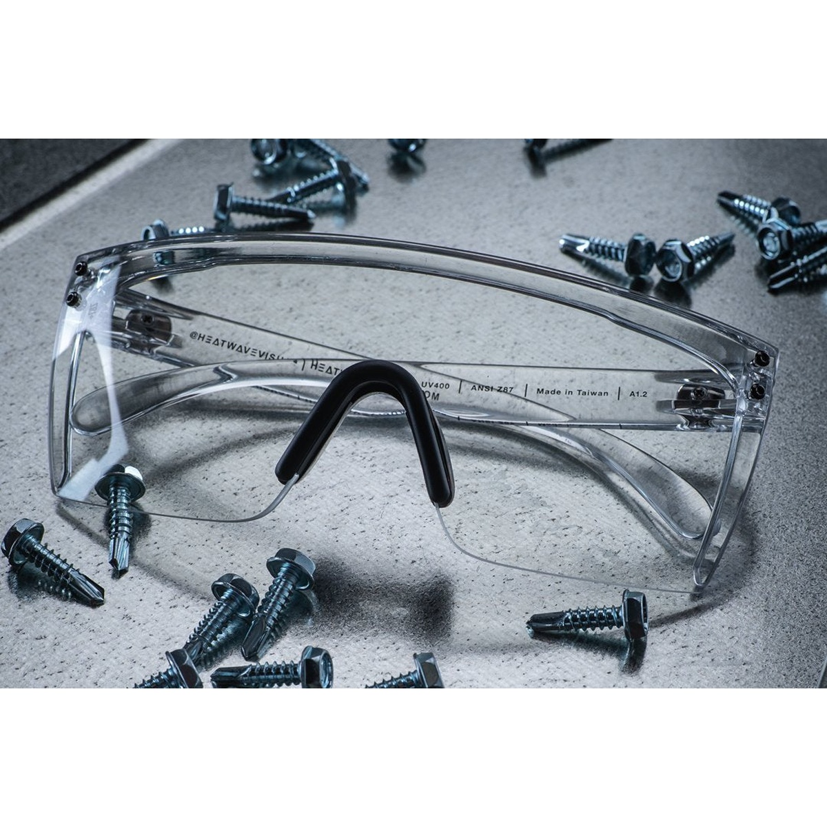 Heat Wave Visual Lazer Face Glasses CLEAR Z87 clear safety glasses on a work surface with metal fasteners, showing wide wraparound lens coverage for shop and jobsite eye protection