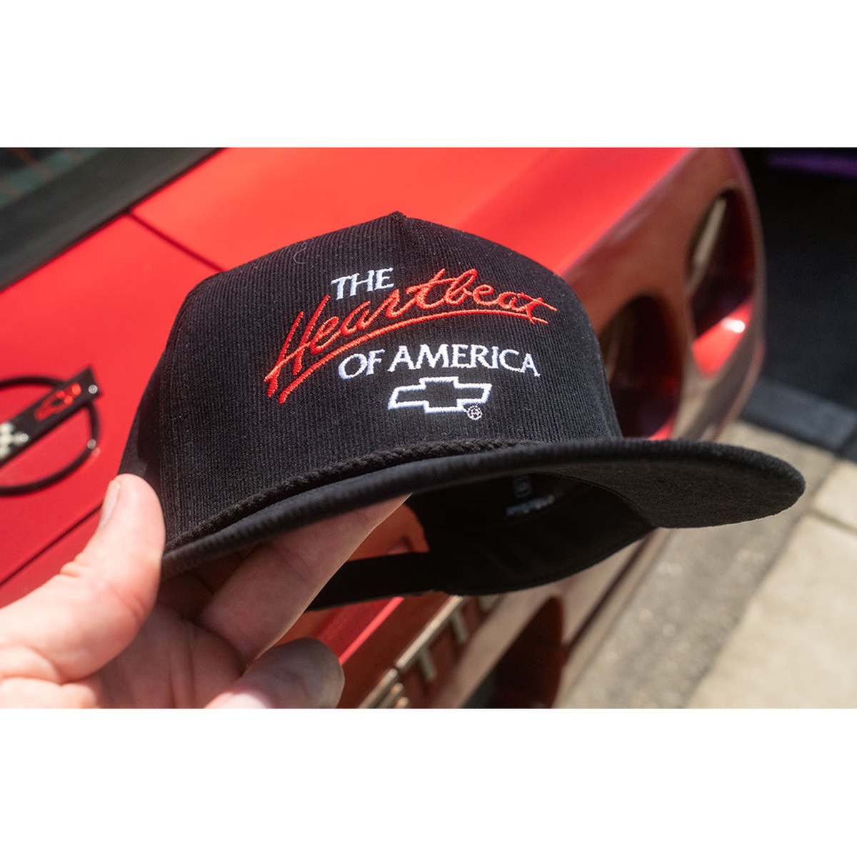 HWV Chevrolet Heartbeat of America Hat - BLACK Cord held outdoors near a red Chevrolet vehicle, showing the embroidered front logo and curved black corduroy bill in automotive lifestyle use