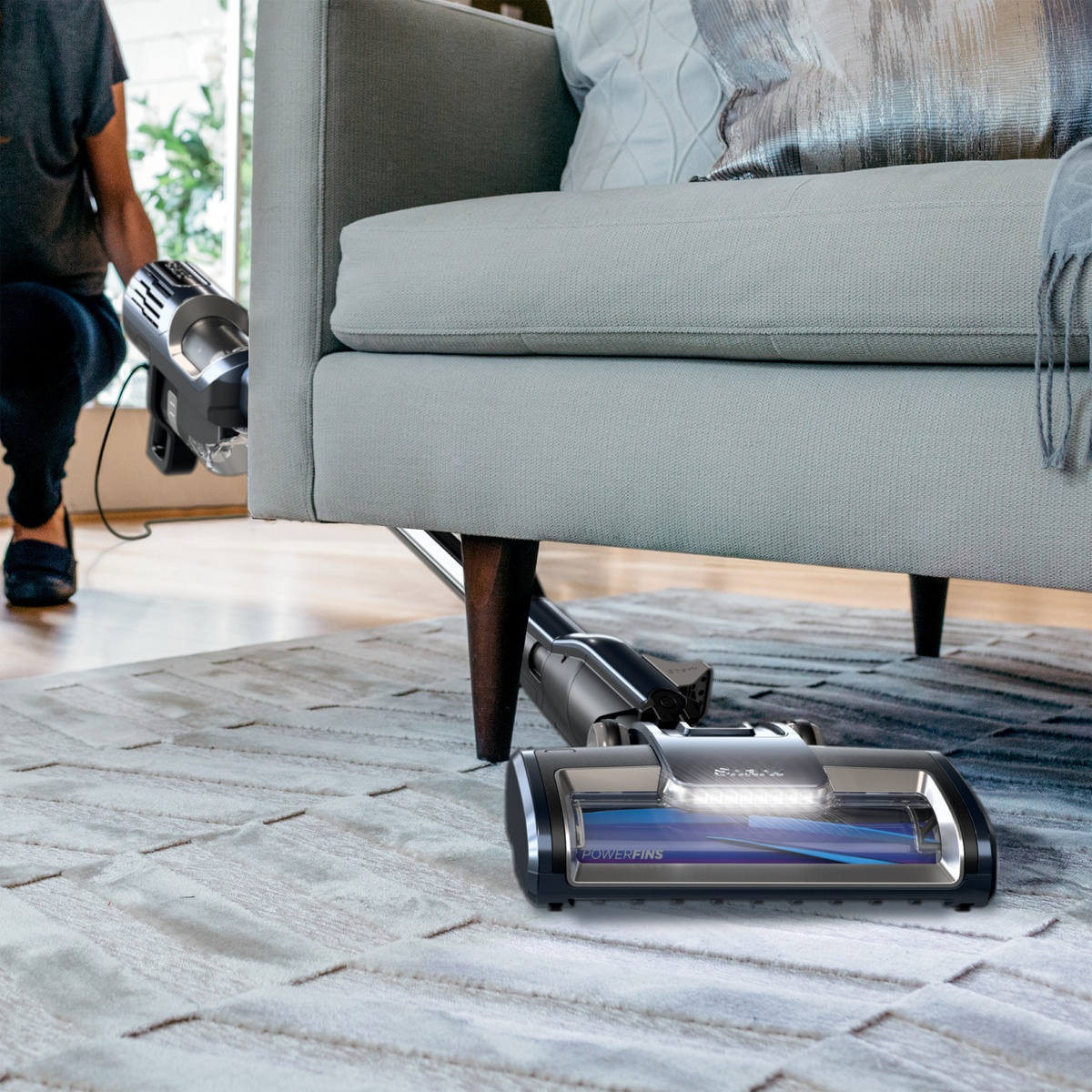 Low-profile floor nozzle with LED headlights reaching under a sofa.