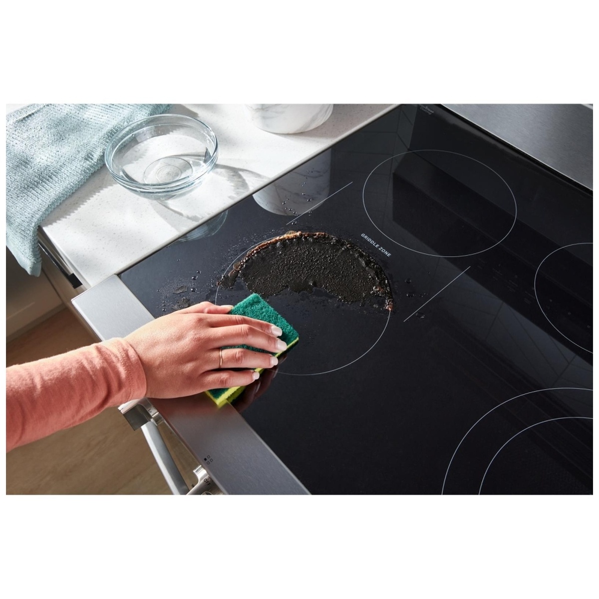 Hand wiping residue from a smooth black glass cooktop with water and a sponge.