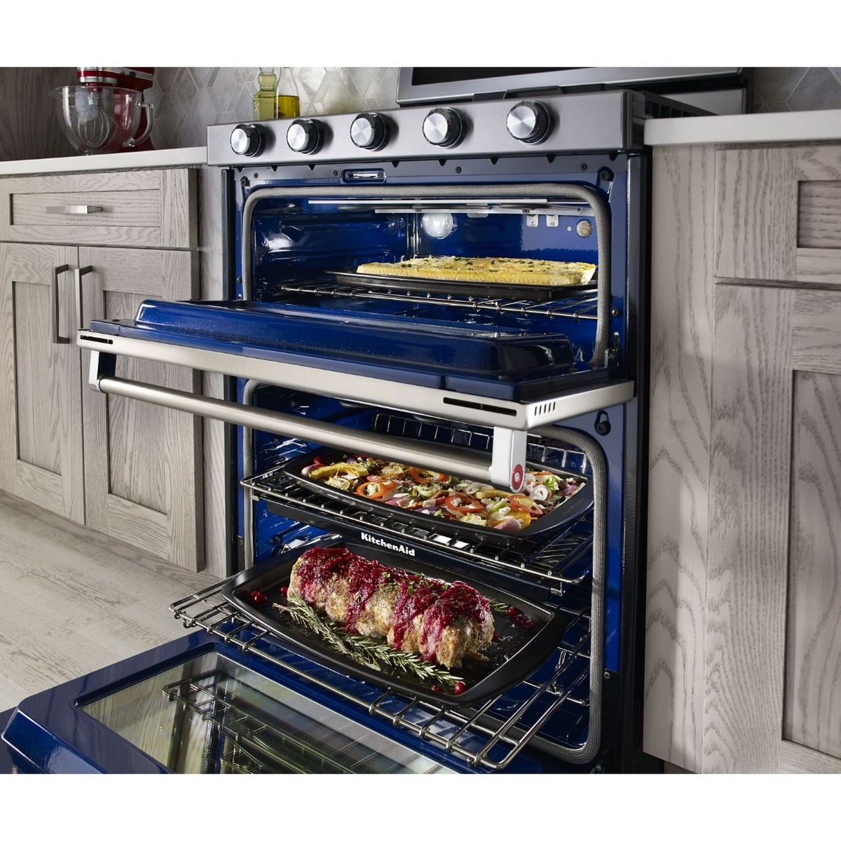 KitchenAid freestanding double oven range with both oven doors open, showing multiple racks and a roll-out rack holding a roasting pan