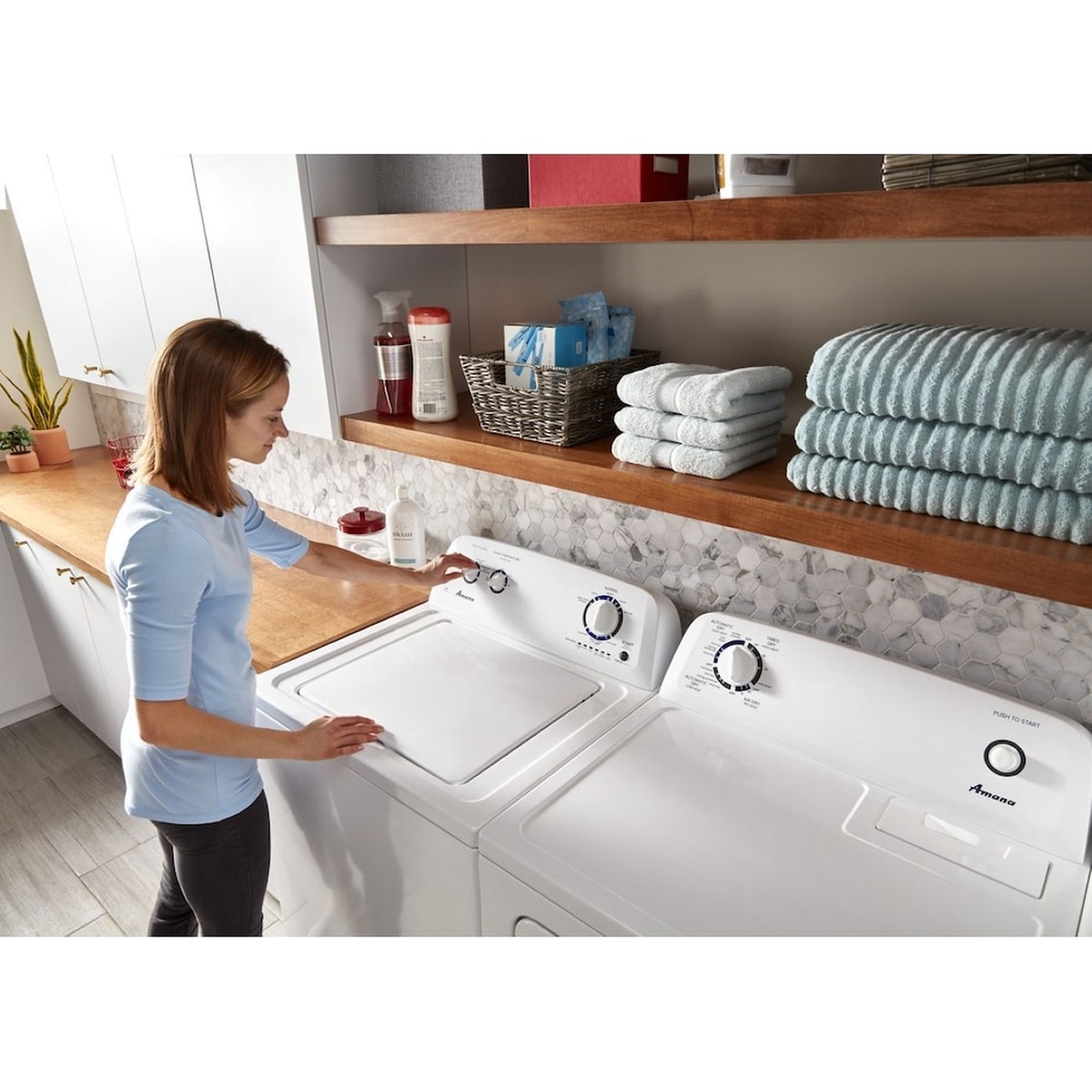 Laundry room lifestyle scene with a person using matching Amana washer and dryer installed side by side under open shelves with folded towels