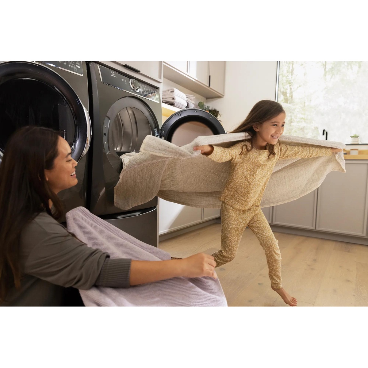 Electrolux front-load laundry pair in a bright laundry room with a child holding a large sheet